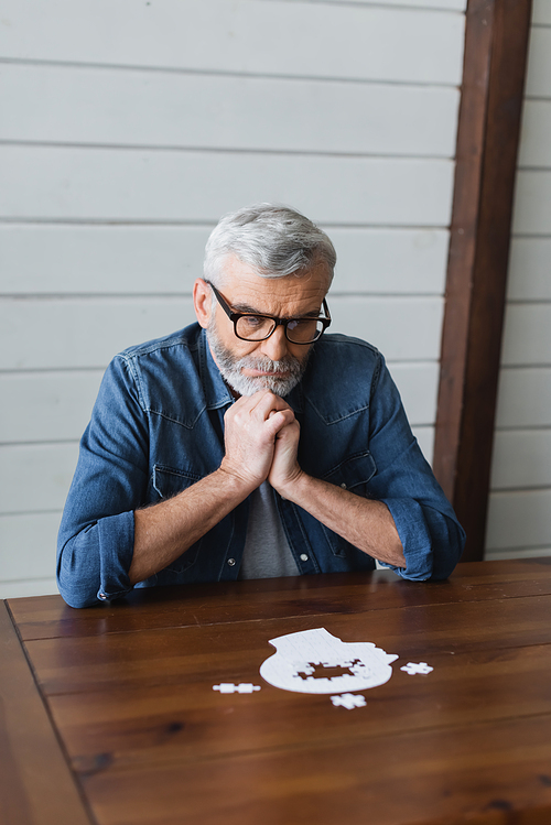 Thoughtful elderly man in eyeglasses looking at blurred puzzle on table