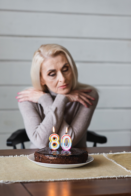 Birthday cake with candles in shape of eighty numbers on table near blurred senior woman