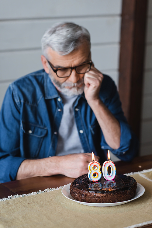 Birthday cake with candles near lonely elderly man on blurred background