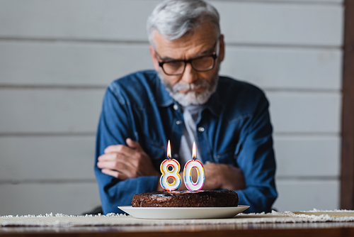Birthday cake with burning candles in shape of eighty numbers near senior man on blurred background