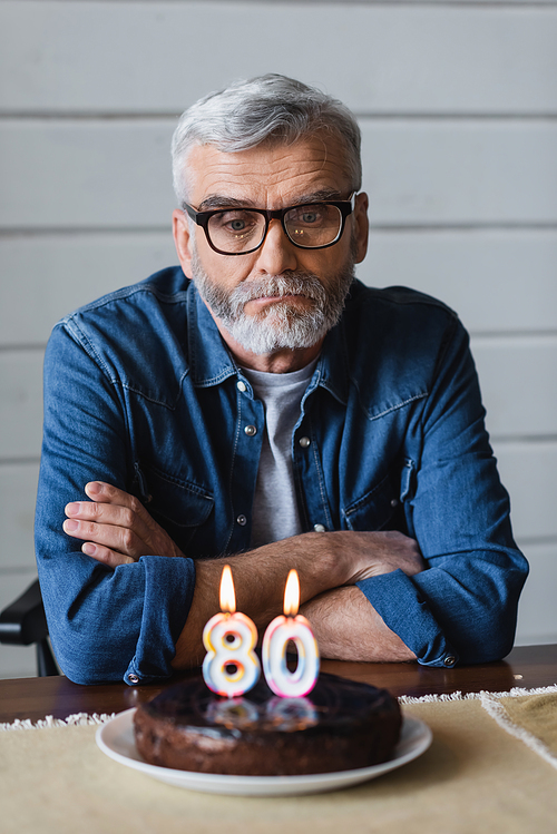 Lonely man in eyeglasses sitting near blurred birthday cake with candles in shape of eighty numbers