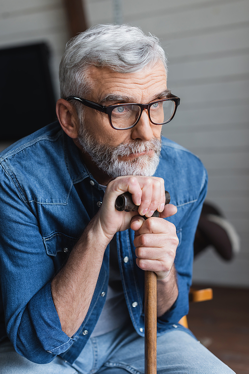 Elderly man in eyeglasses holding crutch at home