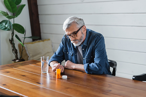 Lonely senior man holding pill near water on table