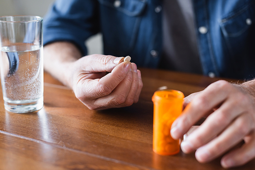 Cropped view of senior man holding pill near jar and glass of water