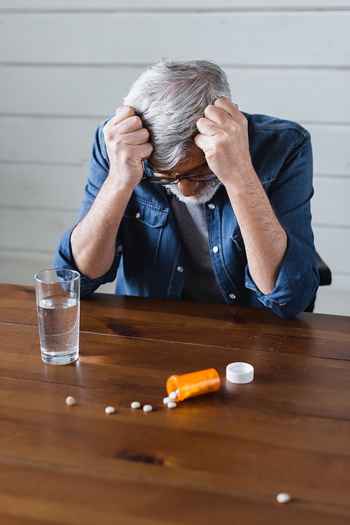 Depressed man looking at blurred pills near glass of water on table