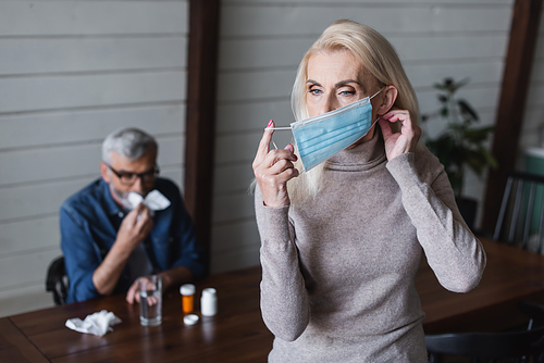 Woman wearing medical mask near blurred husband with napkin and pills