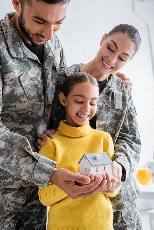 Smiling parents in military uniform holding model of house near child at home