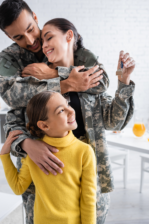 Smiling kid looking at key near parents in military uniform hugging at home