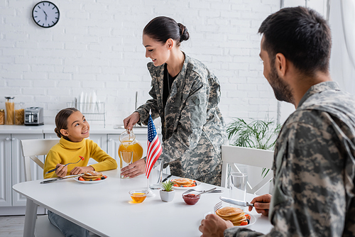 Smiling woman in military uniform pouring orange juice near family and american flag on table at home