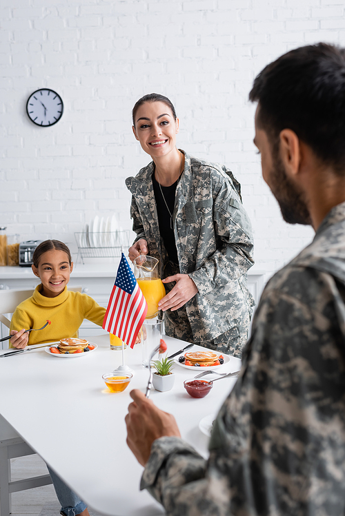 Smiling woman in military uniform holding orange juice near daughter and husband during breakfast at home