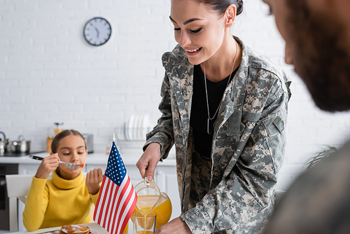 Smiling woman in military uniform pouring orange juice near blurred family and american flag at home