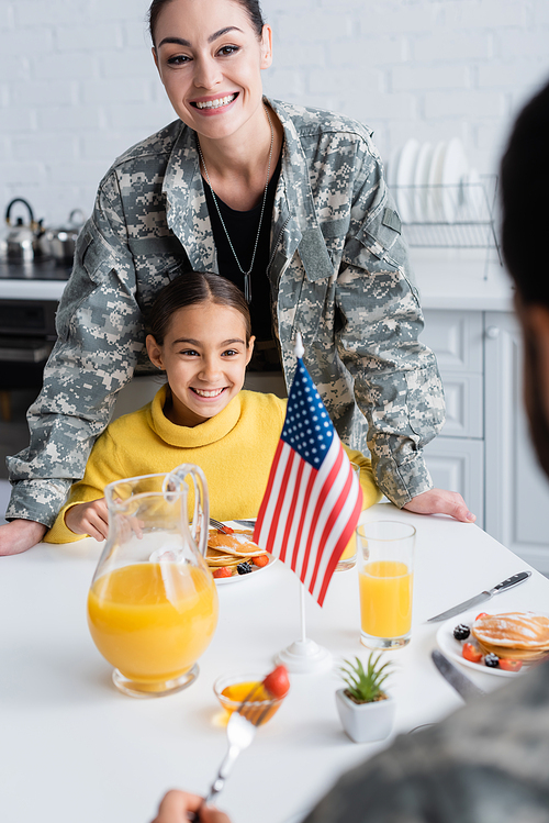 Smiling woman in military uniform standing near daughter, breakfast and american flag in kitchen