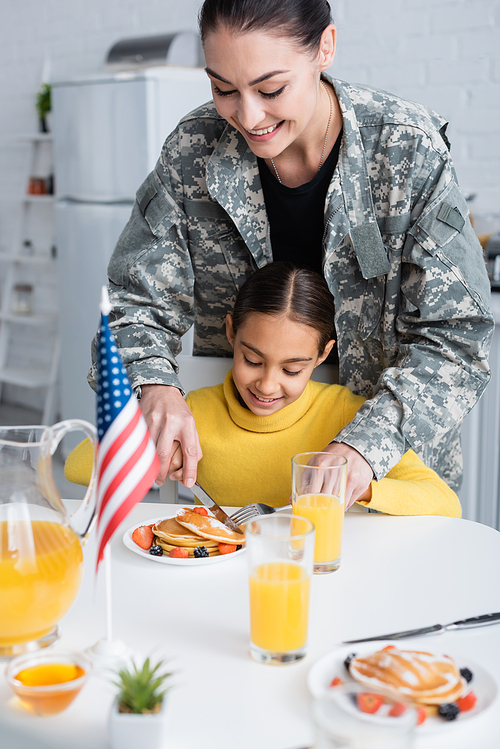 Smiling woman in camouflage uniform cutting pancakes near daughter and american flag in kitchen