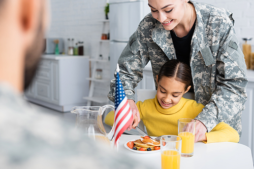 Smiling mother in camouflage cutting pancakes near daughter and blurred husband at home