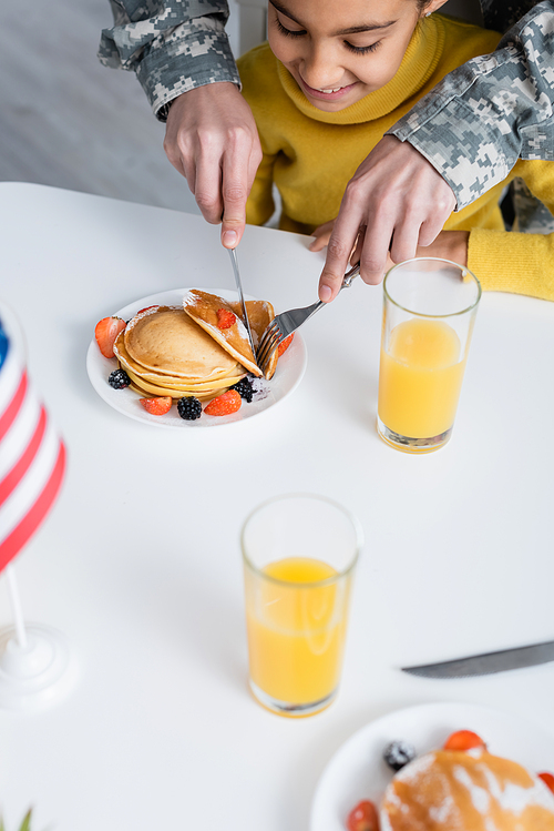 Mother in military camouflage cutting pancakes near smiling daughter and american flag at home