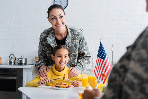 Smiling mother in camouflage uniform standing near daughter and american flag during breakfast in kitchen