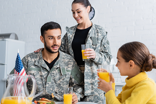 Smiling parents in military uniform looking at daughter near american flag during breakfast at home