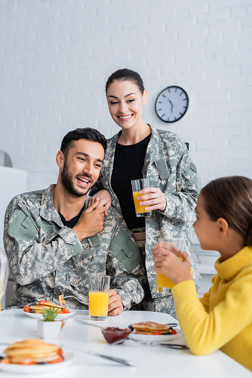 Cheerful parents in military uniform holding hands near daughter during breakfast at home