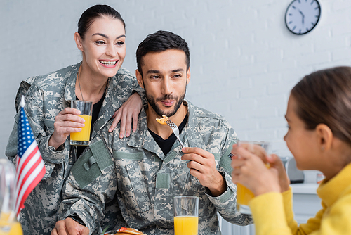 Smiling parents in military uniform looking at child near american flag during breakfast at home