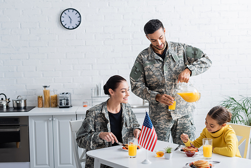 Man in military uniform pouring orange juice near family and american flag in kitchen