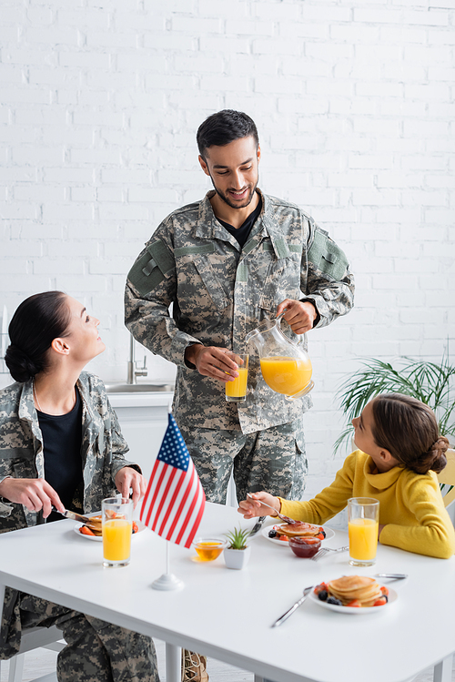 Man in military uniform pouring orange juice near family and american flag during breakfast at home