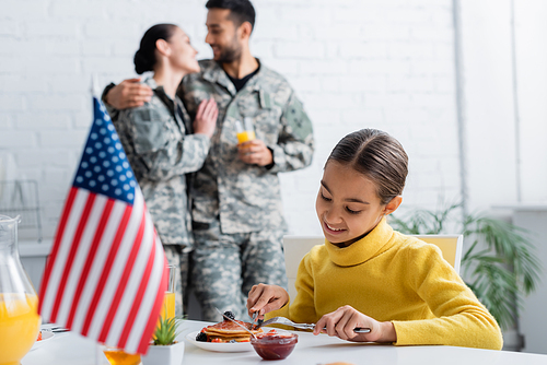 Smiling kid sitting near pancakes, blurred american flag and parents in military uniform at home