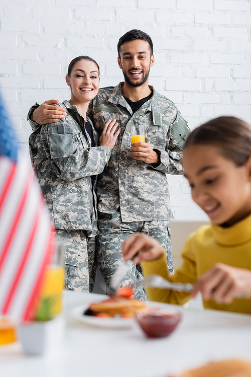 Cheerful parents in camouflage uniform looking at daughter having breakfast near blurred american flag at home