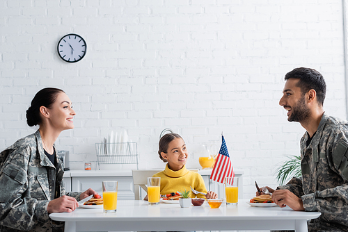Cheerful parents in camouflage sitting near breakfast, american flag and daughter at home