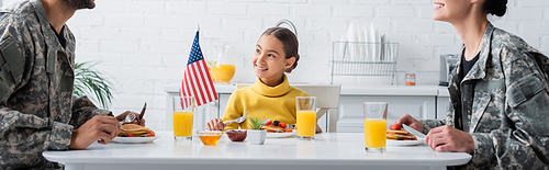 Smiling girl sitting near breakfast, american flag and parents in camouflage, banner