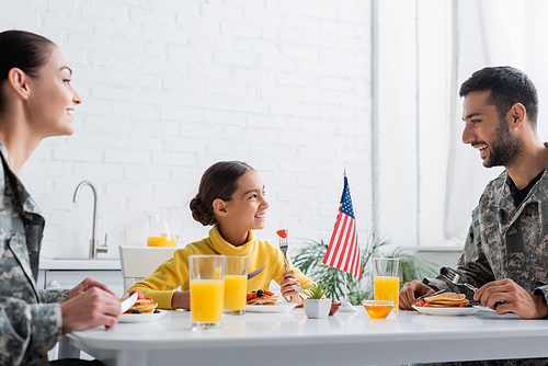 Positive kid looking at parents in military uniform near tasty breakfast and american flag at home