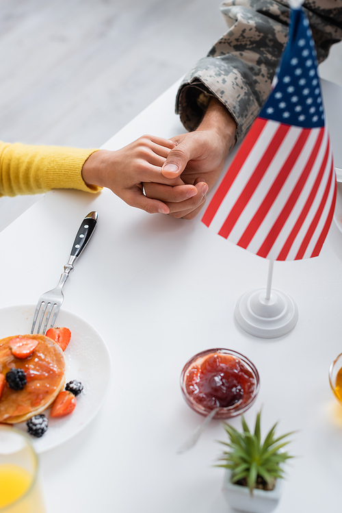 Cropped view of man in military uniform holding hand of daughter near breakfast and american flag