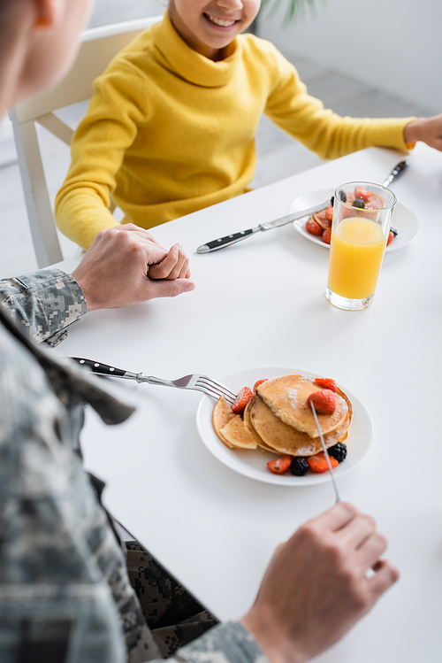Cropped view of woman in military uniform holding hand of smiling child near pancakes and orange juice at home
