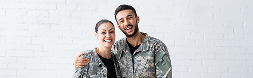 Smiling man in camouflage uniform hugging wife at home, banner