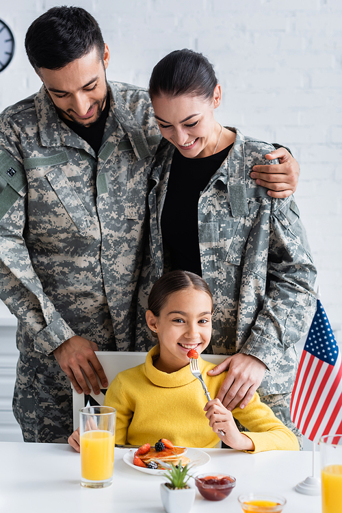 Happy parents in military uniform standing near daughter with breakfast and american flag in kitchen