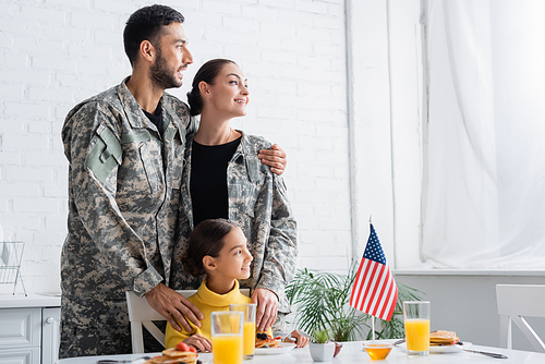 Cheerful parents in camouflage standing near daughter and american flag during breakfast at home