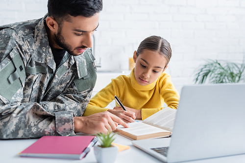 Father in military uniform pointing at book near daughter and laptop at home