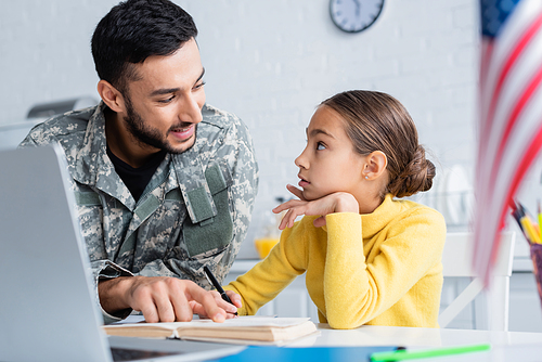 Smiling father in camouflage uniform pointing at book near daughter, laptop and american flag at home