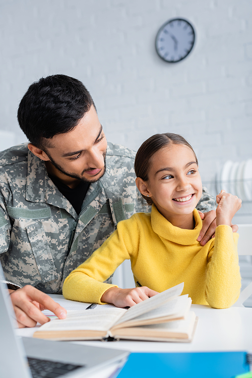 Smiling man in camouflage uniform hugging daughter near book and laptop at home
