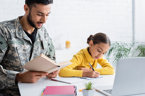 Father in military uniform holding book near daughter writing on notebook and laptop at home