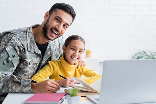 Cheerful man in military uniform hugging daughter near notebooks and laptop at home