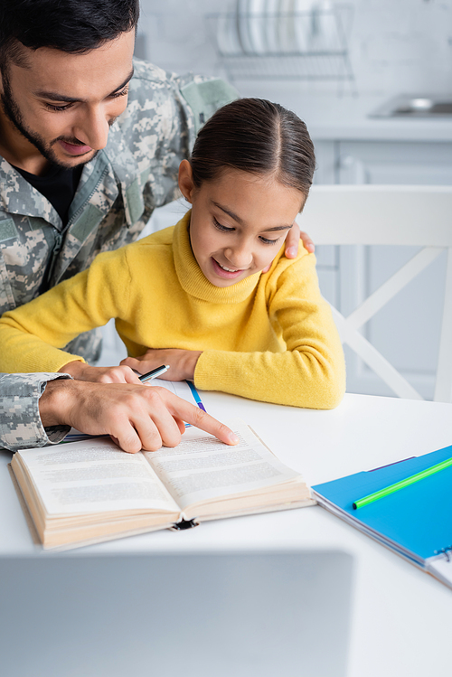 Man in camouflage uniform pointing at book near smiling daughter and laptop at home