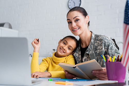 Smiling woman in military uniform holding book near child, laptop and american flag at home