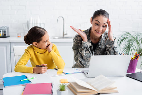 Smiling mother in camouflage uniform looking at laptop near daughter and notebooks at home