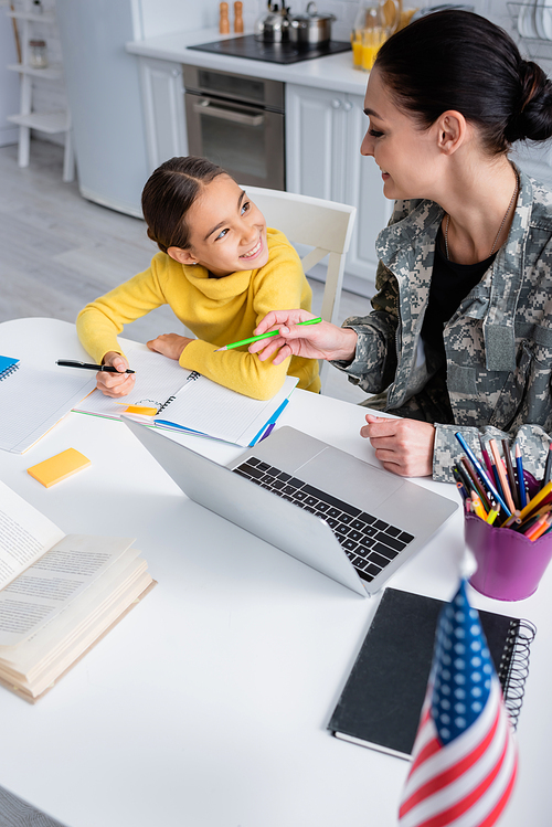 Smiling mother in military uniform holding pencil near daughter with notebook and laptop at home