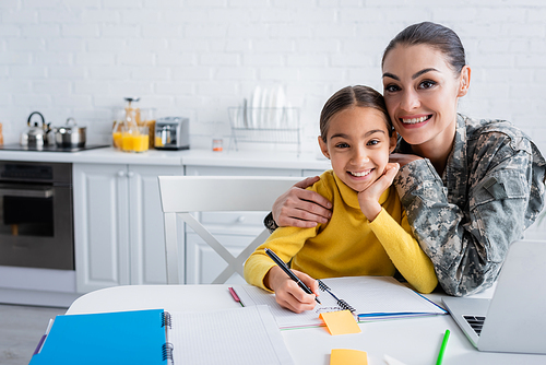 Smiling woman in military uniform hugging child near notebooks and laptop at home