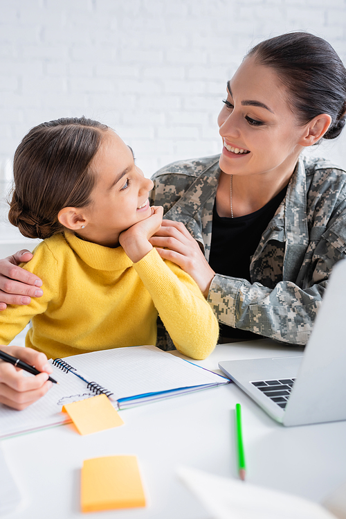 Smiling mother in military uniform looking at kid during schoolwork near laptop at home