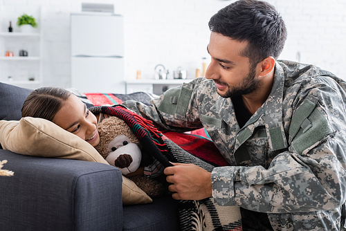 Smiling father in military uniform covering smiling daughter with soft toy on couch at home