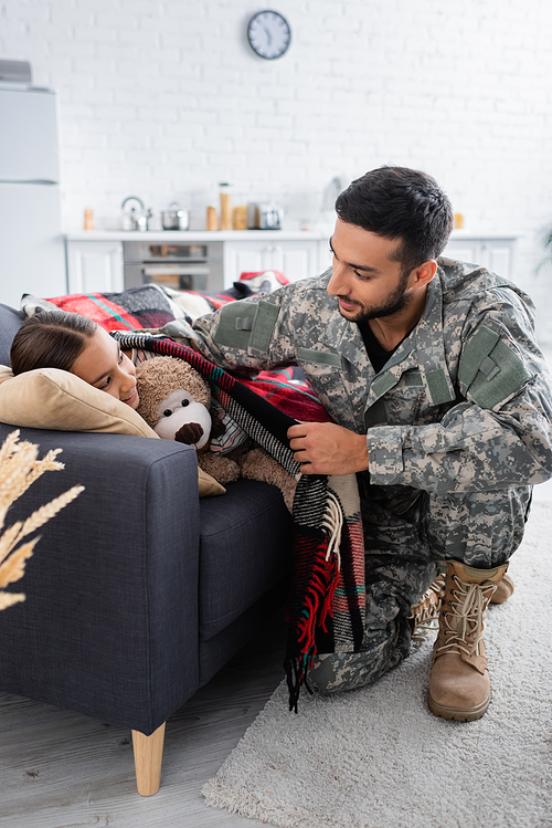 Man in camouflage uniform putting blanket on child with teddy bear on couch
