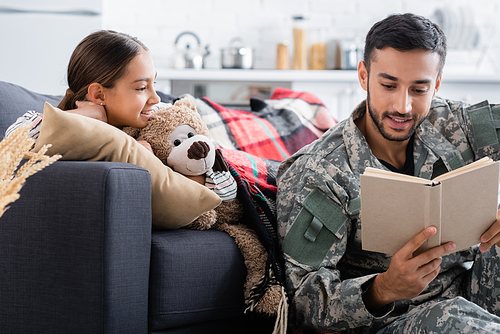 Dad in military uniform reading book near cheerful kid with teddy bear on couch