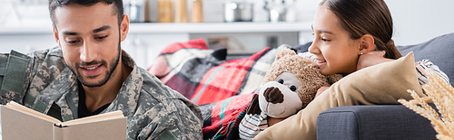 Smiling man in military uniform reading book near child with teddy bear on couch, banner
